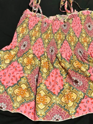 Mönstrad rosa kortklänning med knytband - Färgglad kortklänning med knytband på axlarna och smock upptill. Klänningen har ett unikt mönster i rosa, gult och beige med inslag av blommor och geometriska former. Perfekt för sommarens varma dagar och har en luftig passform.