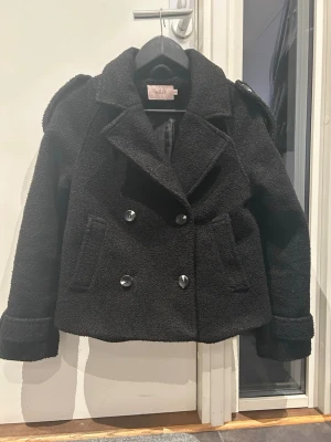 Svart teddykappa från Nelly - Säljer en svart teddyjacka från Nelly med dubbelknäppning och bred krage. Jackan har två framfickor, axelklaffar och är gjord i mjukt, fluffigt material som ger en cool look. Perfekt för dig som vill ha en trendig och mysig jacka. En knapp är lite lös som man kan se på sista bilden men inget som syns när den är stängd och går att sy fast så att den sitter lite bättre, om man skulle vilja det. Säljer pågrund av att den blivit för liten och såklart är jackan tvättad