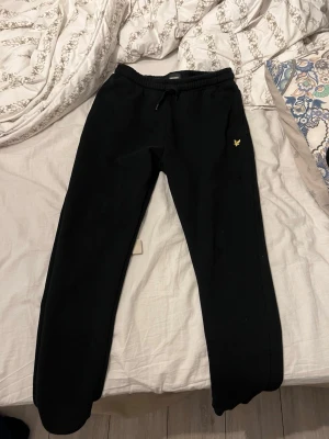 Svarta mjukisbyxor från Lyle & Scott - Svarta mjukisbyxor från Lyle & Scott med resår i midjan och dragsko. Klassisk passform med ribbade muddar vid bensluten och en liten gul logga på vänster lår. Mjukt material, perfekt för chill dagar.