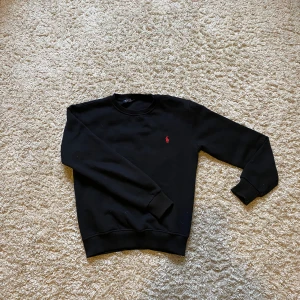 Svart tröja från Polo Ralph Lauren - Svart Polo Ralph Lauren sweatshirt med klassisk röd broderad logga på bröstet. Perfekt basic-plagg med snygg passform och stilren look.