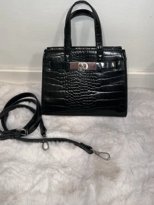 Svart handväska i krokodilimitation - Stilren svart handväska från GinaTricot i glansigt konstläder med krokodilpräglad yta. Väskan har två handtag, avtagbar axelrem och silverfärgade metalldetaljer. Klassisk rektangulär form och vridlås framtill. Perfekt för dig som gillar trendiga accessoarer.