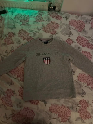 Grå sweatshirt från GANT strl S - Snygg grå sweatshirt från GANT med broderad logotyp och sköld med stjärnor och ränder på bröstet. Tröjan har rund halsringning och långa ärmar. Perfekt för en avslappnad och stilren look.