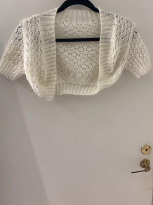 Vit stickad bolero med kort ärm - Söt vit stickad bolero med korta ärmar och öppet framstycke. Tröjan har ett luftigt hålmönster och bred ribbad kant runt hals och nederkant. Perfekt att slänga över en topp för extra stilpoäng.