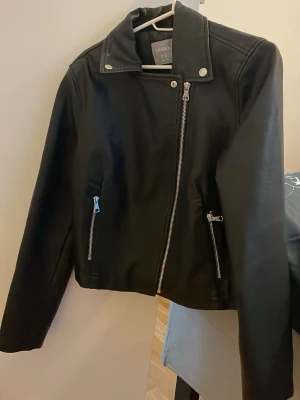 Svart skinnjacka från lindex  - Snygg svart skinnjacka från lindex med klassisk bikerstil. Jackan har sneddragkedja, två fickor med dragkedja framtill och coola detaljer vid axlar och krage. Perfekt för dig som vill ha en stilren och tidlös look.