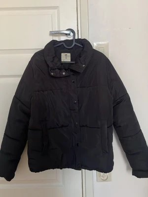 Svart pufferjacka från H&M - Säljer en svart pufferjacka från H&M med hög krage och tryckknappar framtill. Jackan har en boxig passform och två stora fickor på sidorna. Perfekt för kalla vinterdagar och riktigt skön att bära. Knappt använd så den är i mycket bra skick