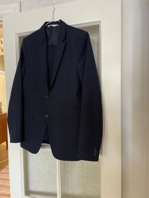 Mörkblå kostym stl 170 - Snygg mörkblå kostym, kavaj med två knappar framtill och klassiska slag. Kavajen har två fickor med lock och knappar vid ärmsluten. Perfekt för en stilren och tidlös look. Passar till både skjorta och t-shirt.