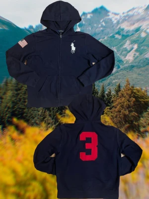 Marinblå zip hoodie Polo Ralph Lauren - Marinblå hoodie med dragkedja från Polo Ralph Lauren. Storlek 14-16 år, passar någon runt 160cm lång. Stor vit logga på bröstet, amerikansk flagga på ena ärmen och stor röd siffra 3 på ryggen. Klassisk huva och kängurufickor framtill. Tillverkad i mjukt bomullsmaterial.
