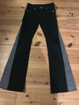 Svarta utsvängda jeans med grå panel - Unika svarta jeans med breda grå paneler längs sidorna och utsvängda ben. Klassisk femficksmodell med två bakfickor som har lock och knapp. Säljer dem på grund av att de inte har kommit till användning, utan bara använts 2 gånger!💓