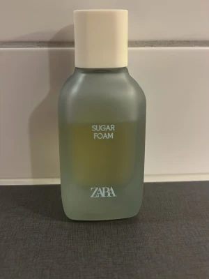 ZARA Sugar Foam parfym - ZARA Sugar Foam parfym i en stilren, frostad glasflaska med vit kork. Flaskan har en mjuk, rektangulär form och minimalistisk design. Perfekt för dig som gillar söta och fräscha dofter.