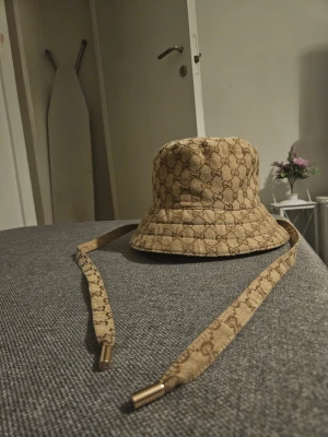 Gucci beige bucket hat med logga - Snygg bucket hat från Gucci i beige med klassiskt GG-monogram över hela hatten. Hatten har bred brätte och långa snören med metalländar för extra stilpoäng. Perfekt accessoar för att lyfta din outfit med lyxig känsla.