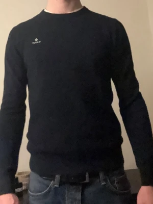 Mörkblå stickad tröja från GANT - Snygg mörkblå stickad tröja från GANT med rund halsringning och diskret logga på bröstet. Tröjan har en klassisk passform och ribbade muddar vid ärmslut och nederkant. Perfekt för lager-på-lager och stilrena outfits.