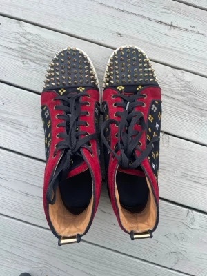 Christian Louboutin röda spikes sneakers - Unika sneakers från Christian Louboutin i röd mocka med svarta detaljer och guldmönster. Tåpartiet är täckt av guldspikar och sidorna har broderade mönster i gult och vitt. Skorna har svarta skosnören och vit sula med gulddetalj vid hälen.