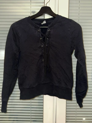 Svart hoodie med snörning fram - Cool svart hoodie med lång ärm och snörning framtill istället för dragkedja eller knappar. Tröjan har ribbade muddar och är tillverkad i mjukt bomullstyg. Perfekt för en avslappnad streetstyle-look.