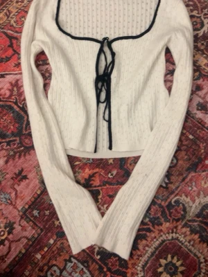 Vit ribbad kofta med svarta detaljer - Trendig vit ribbad kofta med långa ärmar och svart kant längs den fyrkantiga urringningen. Koftan har knytning framtill med svarta band och är figurnära i modellen. Perfekt för lager-på-lager och snygg till jeans. Köpt på New Yorker