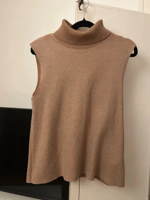 Beige ärmlös polotröja från Zara - Stickad ärmlös polotröja från Zara i en snygg beige nyans. Tröjan har hög, ribbad polokrage och en rak, avslappnad passform. Perfekt att styla över en skjorta eller långärmad topp för en trendig lager-på-lager-look.