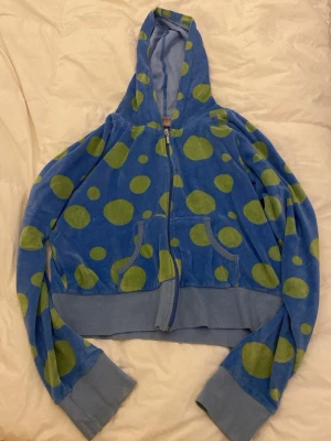 Blå hoodie med gröna prickar - Cool blå hoodie med dragkedja och stora gröna prickar från Stina Kempe. Hoodien har huva, ribbade muddar och fickor framtill. Tillverkad i mjuk frottéliknande material som känns skönt mot huden. Perfekt för en färgglad och lekfull stil.