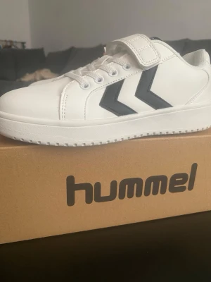 Vita Hummel sneakers med kardborre - Fräscha vita sneakers från Hummel med svarta detaljer och klassiska pilar på sidan. Skorna har både snörning och kardborreband för enkel påtagning. Tillverkade i syntetmaterial med vit sula och Hummel-logga bak.