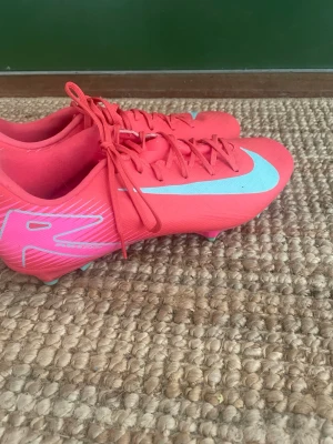 Nike Mercurial rosa fotbollsskor - Säljer ett par Nike Mercurial fotbollsskor i en snygg rosa färg med vita detaljer och logga. Skorna har snörning och är designade för att ge bra grepp på planen. Perfekta för dig som vill sticka ut och ha full kontroll på bollen.