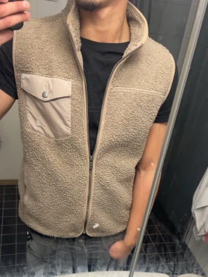 Beige teddyväst från bondelid - Säljer en beige teddyväst från Bondelid med dragkedja framtill, hög krage och kontrasterande bröstficka med lock. Västen har två sidofickor och är gjord i mjukt, fluffigt material som ger en avslappnad och trendig look.