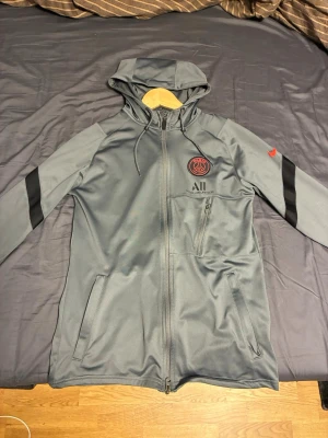 Grå PSG hoodie med dragkedja - Snygg grå hoodie från Paris Saint-Germain med svart och rött klubbmärke på bröstet och 'ALL Accor Live Limitless' tryck. Jackan har hel dragkedja, huva med dragsko och svarta detaljer på ärmarna. Perfekt för fotbollsfans som vill ha en sportig look.