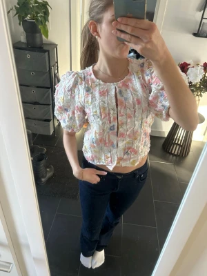 Blommig strukturerad blus från Gina Tricot - Supersöt blus från Gina Tricot med puffärmar och strukturerat tyg. Blusen har ett blommigt mönster i pastellfärger som rosa, blått och gult på vit botten. Kortare modell med rund halsringning och knappar framtill. Perfekt för en trendig look!