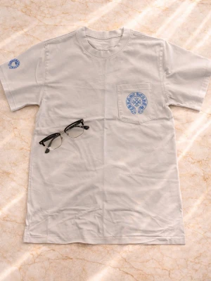 Vit Chrome Hearts t-shirt med blått tryck - Vit t-shirt från Chrome Hearts med blått tryck på bröstfickan och stora blå kors på ryggen. Klassisk passform och rund hals. Tillverkad i mjuk bomull och har korta ärmar. Perfekt för dig som gillar streetwear och snygga detaljer.