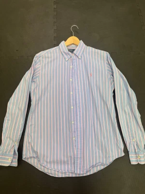 Randig skjorta från Ralph Lauren - Snygg blå skjorta från Ralph Lauren med vita och rosa ränder. Klassisk krage, knäppning framtill och lång ärm. Ikonisk liten logga broderad på bröstet. Perfekt för en stilren look.