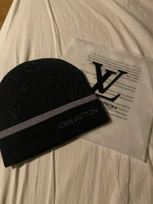 Svart mössa  - Snygg svart mössa med diskret monogrammönster och en grå rand. Märkets logga är broderad framtill. Perfekt accessoar för att lyfta din outfit med en lyxig touch.