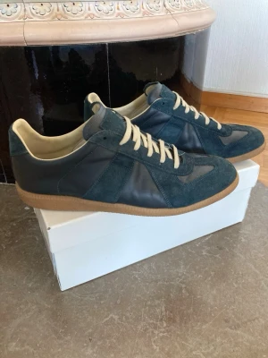 Maison Margiela blå sneakers - Snygga sneakers från Maison Margiela i mörkblå mocka och läder med beige innersula och vita snören. Klassisk gummisula i brunt och diskret branding under sulan. Perfekta för dig som gillar stilrena och exklusiva sneakers.
