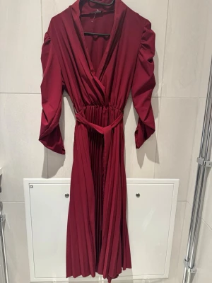 Vinröd omlottklänning med plisserad kjol - Snygg vinröd omlottklänning med plisserad kjol och trekvartsärm. Klänningen har markerad midja med matchande bälte och puffiga axlar för en trendig look. Perfekt för dig som vill sticka ut med stilren elegans.