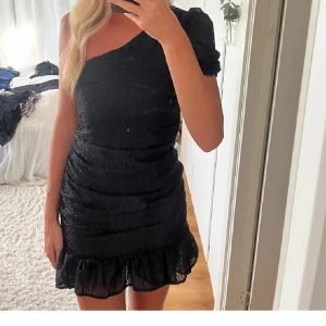 Svart glittrig från vero Moda - Snygg svart kortklänning med glittrigt tyg och oneshoulder-design. Klänningen har volang nertill och sitter tight för en stilren look. Perfekt för dig som vill sticka ut med en trendig och elegant stil.