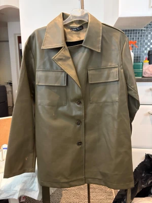 Olivgrön skinnjacka från Weekday - Stilren olivgrön skinnjacka från Weekday med klassisk krage, knäppning framtill och två stora bröstfickor med lock. Jackan har ett avtagbart midjebälte för extra edge och raka ärmar. Perfekt för dig som vill ha en modern look med militärinspirerade detaljer.