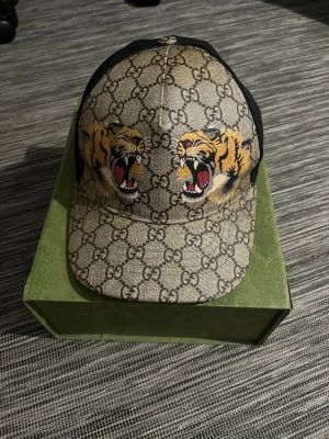 Gucci keps med tigertryck - Säljer en snygg keps från Gucci med klassiskt GG-mönster och två tigrar på framsidan. Beige och svart färgkombination, böjd skärm och mesh på sidorna. Perfekt för dig som vill sticka ut med en exklusiv accessoar. Finns digitalt kvitto som kan vissas. Säljer den akut för behöver pengar. Passar allas huvud om du förstorar eller minskar den
