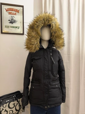 Hollies svart parkajacka med päls - Svart parkas med stor luva kantad av  päls. Jackan har dragkedja, knappar, flera fickor och en figurnära passform. Perfekt för kalla dagar och ger en cool streetstil. Från Hollies Stockholm. 