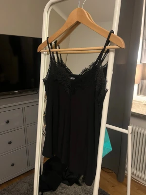 Svart nattlinne med spetsdetaljer - Ett svart nattlinne med tunna axelband och vacker spets längs urringningen. Nattlinnet har en lös och bekväm passform, perfekt för mysiga kvällar hemma. Materialet känns mjukt mot huden och ger en elegant känsla.