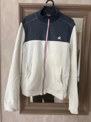 Vit och marinblå hoodie Le Coq Sportif - Snygg hoodie från Le Coq Sportif i vitt och marinblått med dragkedja i rött, vitt och blått. Liten broderad logga på bröstet och två fickor framtill. Mjuk bomullskänsla och hög krage för extra stilpoäng.