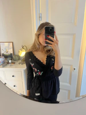 Odd Molly omlottblus - Svart broderad omlottblus med långa ärmar från Odd Molly. Blusen har knytband i midjan och dekorativa spetsdetaljer längs öppningen. I mycket fint skick. storlek 36