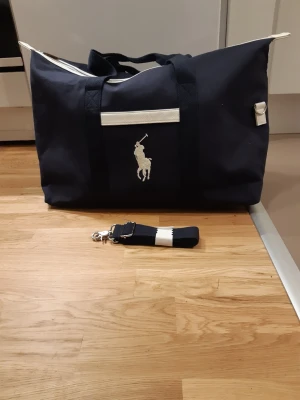 Klassisk Weekendbag från Polo Ralph Lauren – Marinblå - Väskan är tillverkad i slitstark bomullskanvas och har robusta bärhandtag samt en avtagbar axelrem för maximal komfort. Märke: Polo Ralph Lauren Färg: Marinblå med vita detaljer Skick: Aldrig använd Mått: Ca 50 x 30 x 22 cm – rymmer allt du behöver för en övernattning. Övrigt: Dragkedja upptill och rymligt huvudfack.