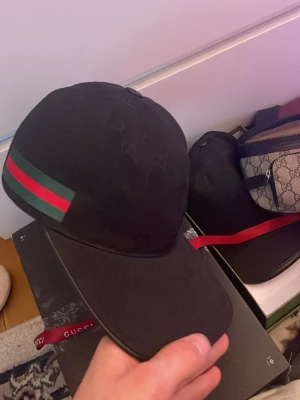 Svart GG keps med GG-mönster - Säljer en svart keps från GG med klassiskt GG-monogram över hela kepsen och den ikoniska gröna och röda randen på sidan. Kepsen har böjd skärm och justerbar rem bak. Materialet är canvas med detaljer i läder. Perfekt för dig som vill ha en stilren och lyxig accessoar.