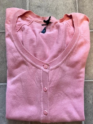 Ljusrosa kofta med knappar - Söt ljusrosa kofta med rund halsringning och knäppning framtill. Koftan har två små fickor framtill och ribbade muddar vid ärmslut och nederkant. Perfekt att slänga över en topp för en chill look.