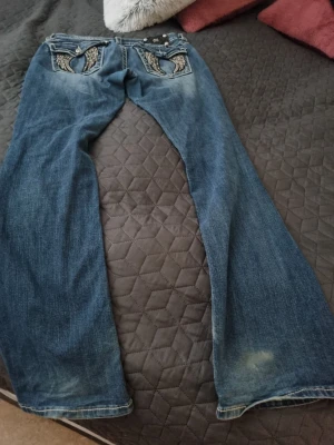 Blå bootcut jeans med broderi från miss me  - Säljer ett par blå bootcut jeans med snygga slitningar och broderade detaljer på bakfickorna. Jeansen har låg midja och kontrastsömmar som ger en cool look. Perfekta för dig som gillar en avslappnad men trendig stil. Storlek 31 använd två gånger 