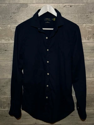 Mörkblå skjorta från Polo Ralph Lauren - Klassisk mörkblå skjorta från Polo Ralph Lauren med vita knappar och lång ärm. Skjortan har en stilren design och är tillverkad i bomull, perfekt för en clean och tidlös look.