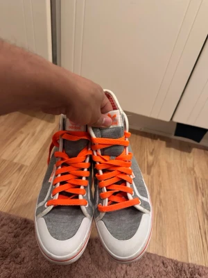 Adidas grå sneakers med orange detaljer - Grå sneakers från Adidas med vita och orange detaljer samt orange skosnören. Skorna har en klassisk siluett med vit sula och Adidas-logga på plösen och sidan. Materialet är textil med inslag av syntet. Perfekt för dig som vill sticka ut med färgglada detaljer.