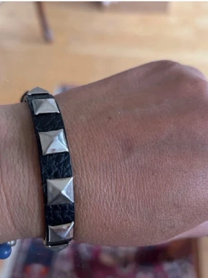 Svart läderarmband med nitar - Snyggt svart armband i läder med silvriga pyramidformade nitar längs hela bandet. Armbandet har ett spänne i metall för justerbar storlek och ger en cool, rockig vibe till din outfit.