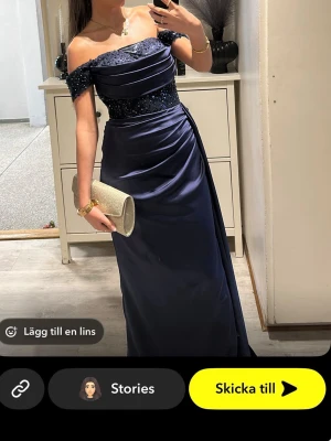 Marinblå offshoulder balklänning - Elegant marinblå långklänning med offshoulder-design och glittriga paljettdetaljer upptill. Klänningen har draperad midja och slits på sidan för extra stil. Perfekt för bal eller festliga tillfällen. Materialet känns lyxigt och har en fin glans. Super fin till Bal eller bröllop!