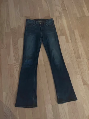 Lågmidjade blå bootcut jeans från Lee - Snygga blå bootcut jeans från Lee med klassisk femficksdesign och gylf med knapp. Jeansen har en mörk tvätt och subtila slitningar för en cool look. Perfekta för dig som gillar retrostil och vill ha ett par jeans med lite vidare ben. De har små defekter längst ner som knappt märks av. Storlek 26/31.
