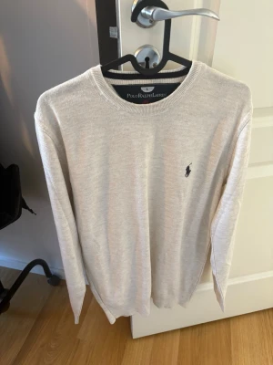 Beige tröja från Polo Ralph Lauren - Snygg beige tröja från Polo Ralph Lauren med rund halsringning och klassisk broderad logga på bröstet. Tröjan har långa ärmar och är tillverkad i ett mjukt material som passar perfekt till många olika outfits.