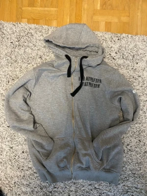 Grå Gymgrossisten hoodie med dragkedja - Säljer en grå hoodie från Gymgrossisten med svart tryck på bröstet och ryggen. Hoodien har dragkedja, två fickor fram och svart snöre i huvan. Tillverkad i mjukt sweatshirtmaterial, perfekt för gymmet eller chill.