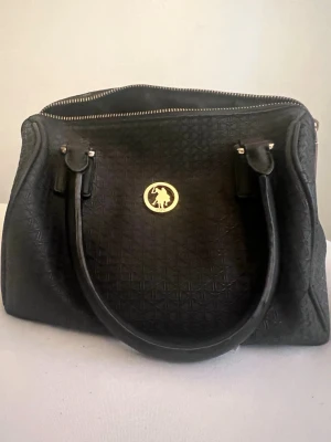 Svart handväska från U.S. Polo Assn. - Snygg svart handväska från U.S. Polo Assn. med guldfärgad logga framtill. Väskan har två rejäla handtag, diskret mönstrad yta och dragkedja upptill. Insidan är fodrad med logotyp-tyg och har innerfack med dragkedja. Perfekt för dig som gillar klassisk och stilren design.