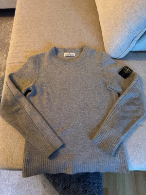 Grå stickad tröja från Stone Island - Snygg grå stickad tröja från Stone Island med klassisk rund hals och ribbade muddar. Tröjan har den ikoniska svarta patchen med gul logga på vänster ärm. Perfekt för dig som gillar stilrena och exklusiva plagg. Svår att hitta, nypris 4899kr en riktigt najs tröja som är 100% äkta ull så den håller fint på kalla dagar, kom ihåg att den sitter lite löst den nere vid händerna inte riktigt så tajt, tröjan har ett hål vid armhålan på höger sida (men inte stort)